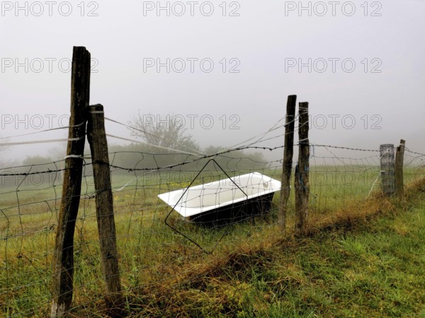 A rusted bathtub rests against a weathered fence, partially hidden by thick fog in a serene rural area during early morning. The mist enhances the peaceful atmosphere. france