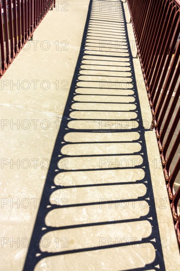 Bright sunlight creates intricate shadows from a red railing onto a stone pathway in a park, highlighting the textures of the ground and the railing's design