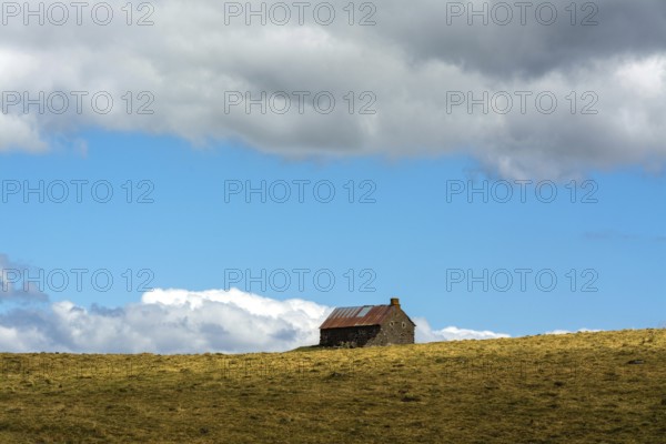 A stone cottage rests on a grassy slope, surrounded by expansive hills. The sky features vibrant clouds and blue patches, creating a serene rural atmosphere. Cezallier. Puy de Dome. Auvergne Rhone Alpes. France