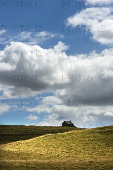 Ruined stone cottage stands alone on a grassy hillside under a partly cloudy sky in a remote countryside setting. Cezallier. Puy de Dome. Auvergne Rhone Alpes. France