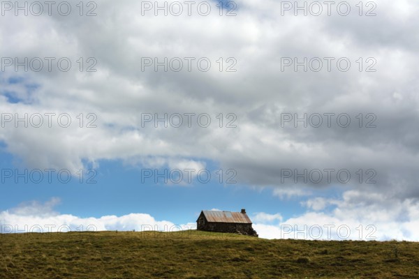 A small cabin sits alone atop a grassy hill, surrounded by wide-open space. The sky is filled with fluffy clouds, creating a serene atmosphere in this rural landscape. Cezallier. Puy de Dome. Auvergne Rhone Alpes. France