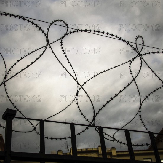 Rusty barbed wire coils twist against a dark and turbulent sky, creating a sense of confinement and unease. The storm clouds loom over a distant structure, emphasizing isolation