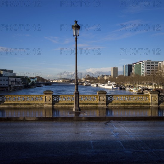 A street light stands tall on a bridge, casting light on the path as the serene Seine River flows beneath. Nearby buildings reflect a calm sky, showcasing urban life in Paris. France