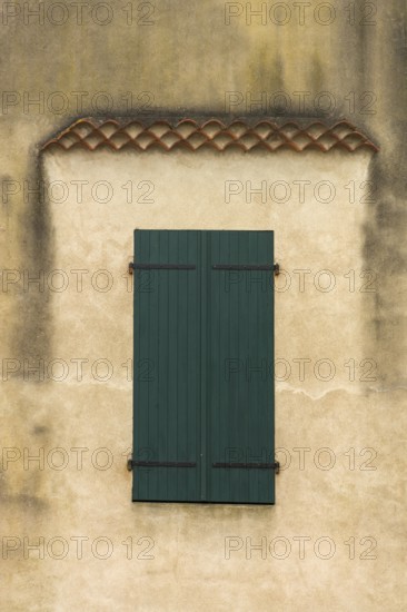 The facade features a pair of blue-green wooden shutters on a weathered wall, showcasing texture and subtle architectural details highlighting the age and character of the house. France