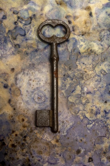 An old key lies against a blue and beige textured backdrop, showcasing its rust and wear. The key's unique shape draws attention, evoking curiosity about its history and purpose
