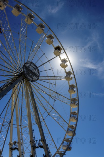 Bright sun shines behind a towering Ferris wheel, creating a vibrant atmosphere at an amusement park. Visitors are drawn to the fun and stunning views offered by the iconic ride