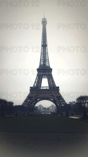 Eiffel Tower stands tall against a grey sky in Paris, surrounded by landscaped gardens. Visitors stroll through the area, enjoying the iconic landmark that symbolizes romance and history. France