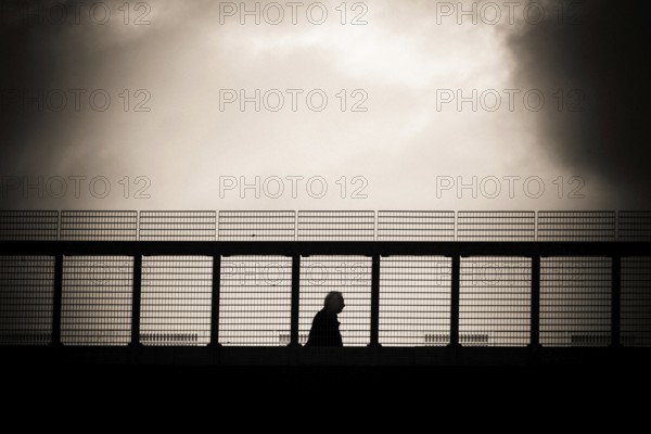 A solitary figure walks along a metal gangway in Lausanne, Switzerland, with a dramatic cloudy sky in the background, capturing a moment of reflection and solitude during late afternoon