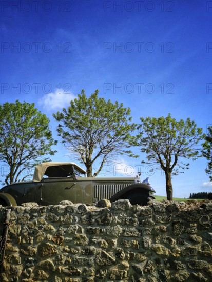 A vintage convertible car sits next to a stone wall, surrounded by lush green trees. The bright blue sky enhances the charming countryside atmosphere