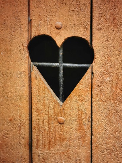 A rustic wooden door features a heart-shaped cutout that reveals iron bars behind it. The warm orange color adds charm to the unique design, enhancing the door's character