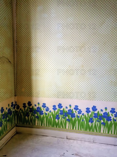 An empty corner room features a unique wall design, showcasing vibrant blue flowers and green grass painted along the base. The artistic touch adds character to the space