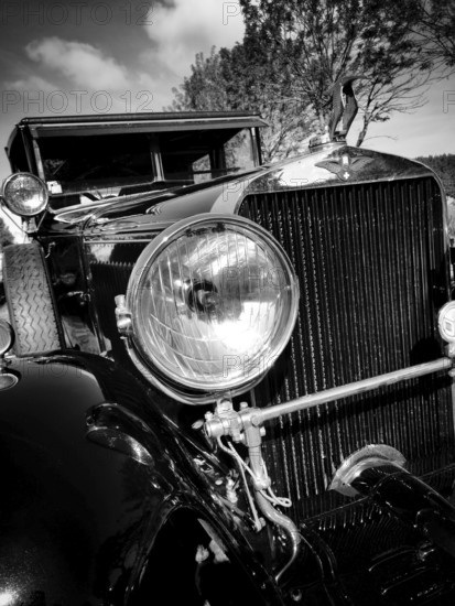 This vintage car displays its polished headlight and elegant grille. The scene captures the beauty of classic automotive design against a bright sky and trees