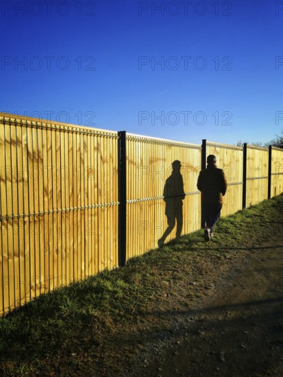 A solitary figure strolls beside a tall wooden fence on a well-worn path. The sun casts elongated shadows, highlighting the serene atmosphere of the late afternoon