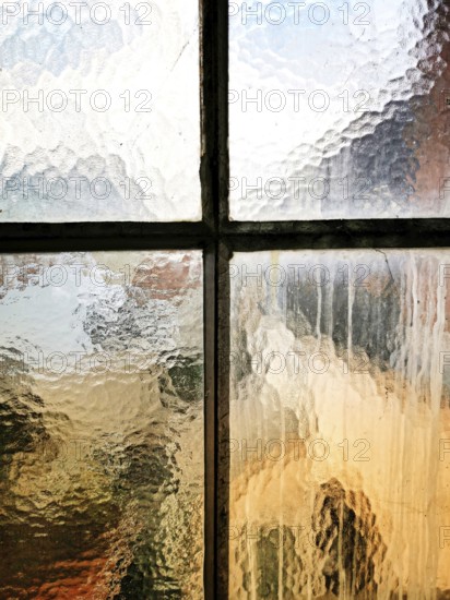 A textured glass window shows raindrops trickling down. The blurred outlines of an urban landscape and warm colors create a cozy, serene atmosphere