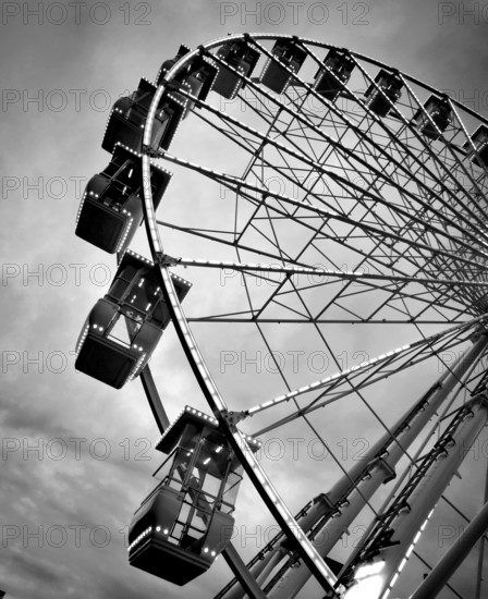 A large Ferris wheel stands prominently against a backdrop of clouds, its illuminated cabins showcasing a fun atmosphere at an amusement park. The evening light creates a dramatic silhouette