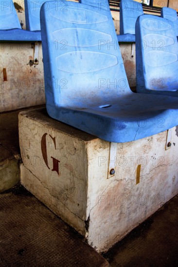 Rows of empty blue seats rest on a weathered concrete base in a deserted sports arena, highlighting the passage of time and lack of maintenance within the neglected venue