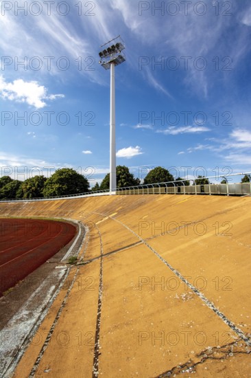 Bright daylight reveals an empty sports stadium with a vibrant red track. The tall floodlights, overgrown with neglect, indicate years without activity. The clear blue sky adds to the desolation. France