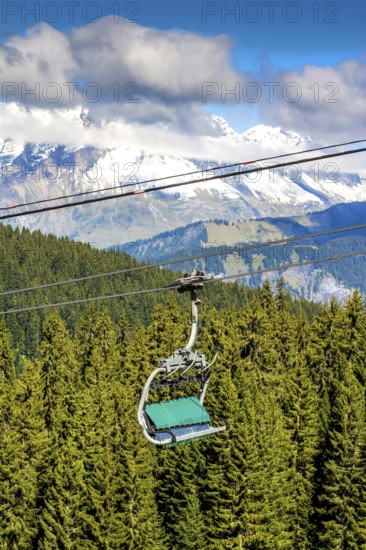 A ski lift transports visitors above green forests toward snowy mountain peaks in Haute-Savoie, France. The clear sky enhances the breathtaking view of the landscape