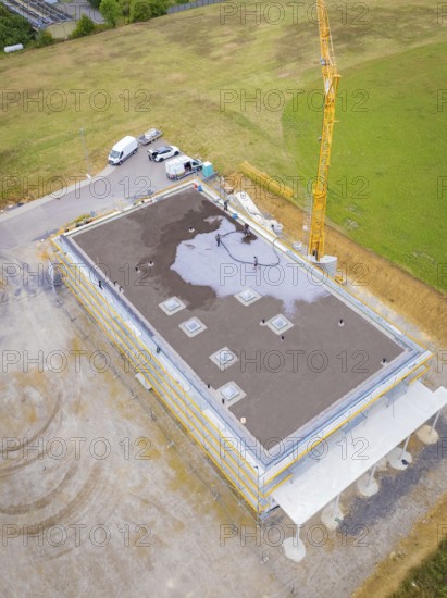 Building in shell construction with crane and vehicles on an open plot with surrounding fields, green roof, house construction, climate neutral