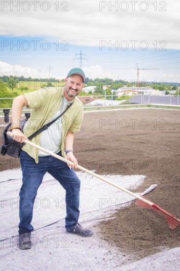Man with rake smiling on a construction site on the roof of a building, roof greening, house construction, climate neutral