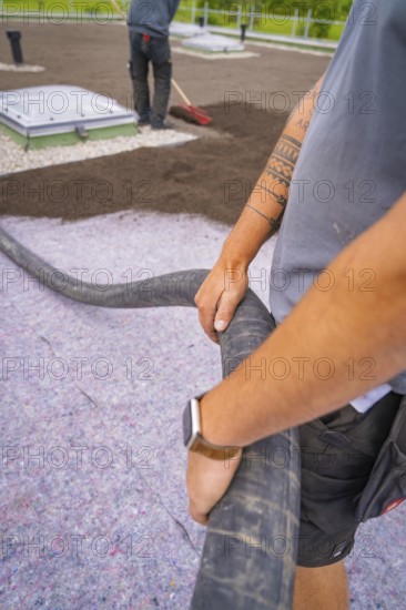 Worker holds a hose with visible tattoos and uses construction clothing, roof greening, house construction, climate neutral