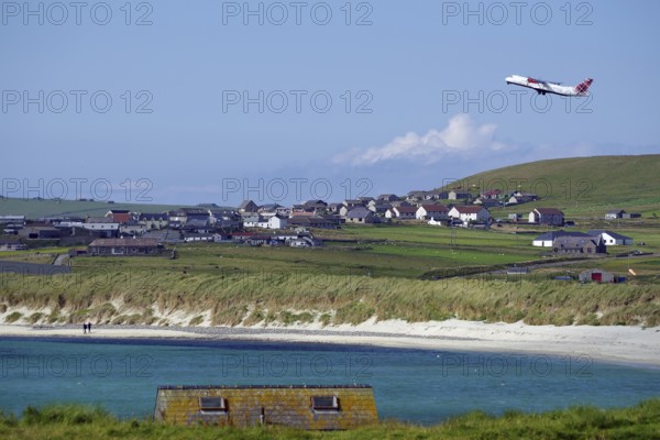 An aeroplane takes off near a rural village with beach and meadows, Sumburgh Head, Shetland Islands, Scotland, United Kingdom