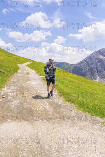 A hiker walks on a path amidst green meadows under a blue sky with clouds, Alpe di Siusi, Dolomites, South Tyrol, Italy