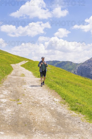 A hiker walks on a path amidst green meadows under a blue sky with clouds, Alpe di Siusi, Dolomites, South Tyrol, Italy