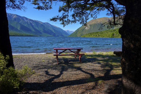 A shaded picnic table on the shore of a lake, with mountains and clear sky in the background, summer, Lake Rotoiti, Saint Arnaud, Tasman Region, South Island, New Zealand