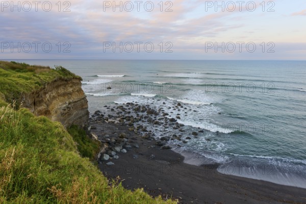 Rocky cliffs by the sea with waves and green vegetation under a cloudy sky, summer, Opunake, Taranaki, North Island, New Zealand
