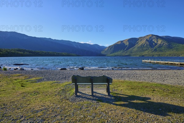 A park bench on the shore of a lake with a view of the surrounding mountains and a boardwalk under a clear blue sky, summer, Lake Rotoiti, Saint Arnaud, Tasman Region, South Island, New Zealand