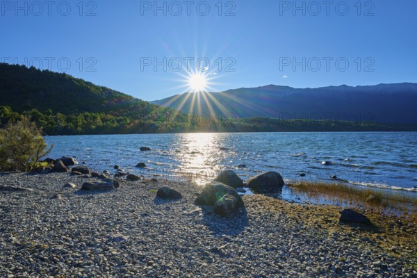 The sun casts dazzling rays over a peaceful lake, framed by rocky shores, summer, Lake Rotoiti, Saint Arnaud, Tasman Region, South Island, New Zealand