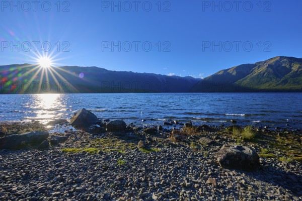 Sunlight dazzles over the horizon as it hits the tranquil lake and surrounding mountains, summer, Lake Rotoiti, Saint Arnaud, Tasman Region, South Island, New Zealand