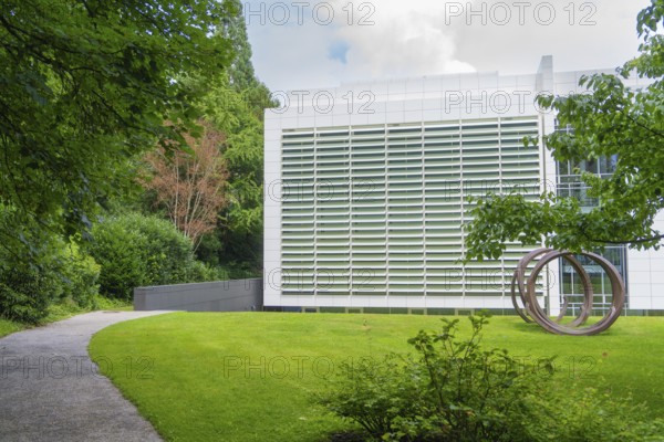 Modern building with a green façade, surrounded by nature and a sculpture in front. A path leads through the grounds, Baden Baden, Germany