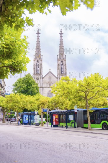 Gothic church with towers behind trees, buses stop on a busy street, Baden Baden, Germany