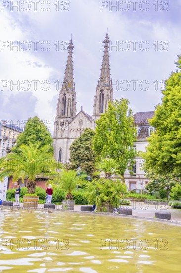 Gothic church with towers in the background, a fountain and palm trees in the foreground, people walking, Baden Baden, Germany