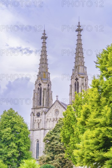 Two tall Gothic church towers rise against a cloudy sky, framed by green trees, Baden Baden, Germany