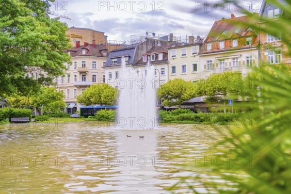 A fountain in an urban pond, surrounded by buildings and green vegetation, Baden Baden, Germany