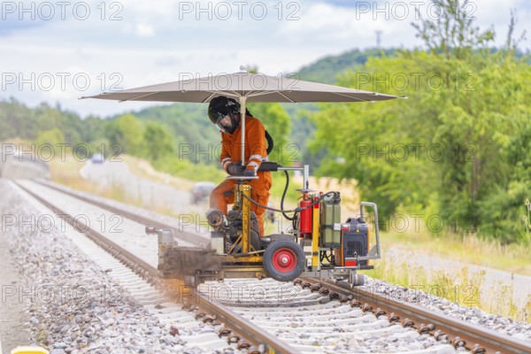Worker in orange overalls operating a machine on railway tracks, protected by a large umbrella, grinding work on the Hermann Hesse railway, Ostelsheim, district of Calw, Germany