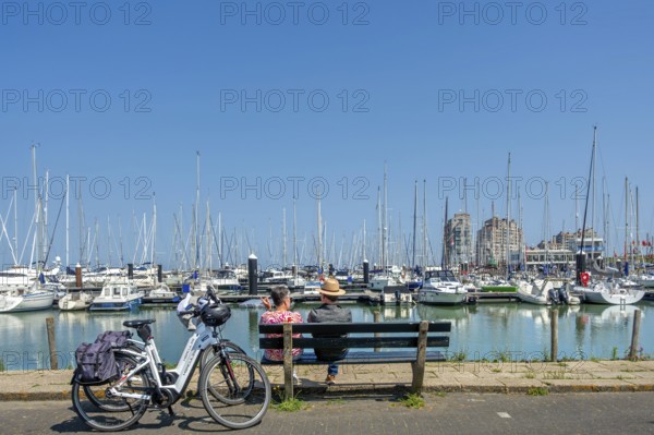Couple of cyclists resting on bench looking over sailing boats in marina of seaside resort Breskens along the Western Scheldt, Zeeland, Netherlands