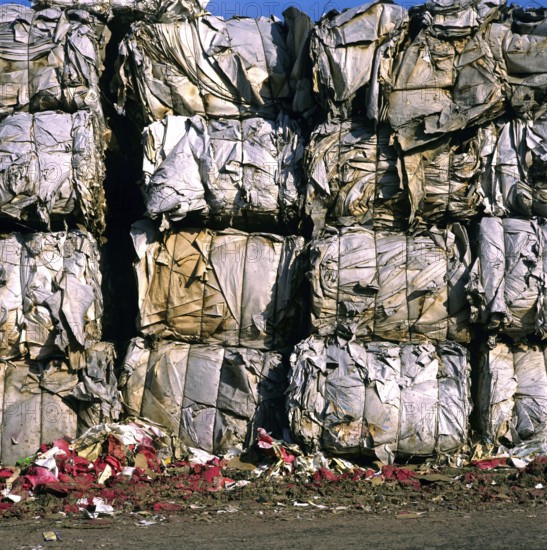 Large compressed bundles of paper products stand stacked at a recycling center, showcasing the effort to recycle waste materials during a sunny day. The surrounding area has scattered debris