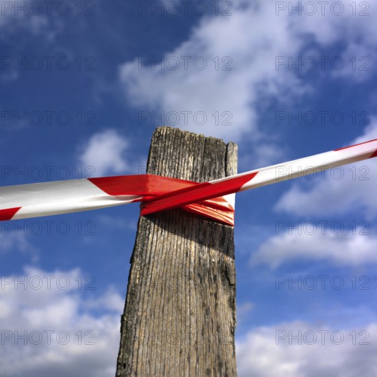 A wooden post stands against a vibrant blue sky, wrapped tightly with red and white barrier tape indicating restricted access. Fluffy clouds add depth to the sky