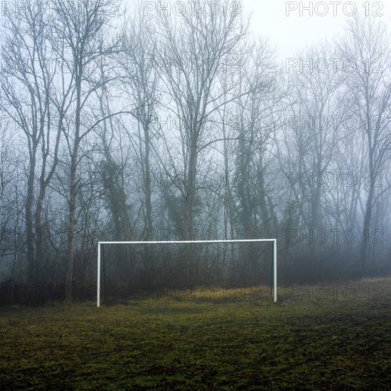 A soccer field shrouded in thick fog features a white goalpost in the foreground. Leafless trees loom in the background, creating an eerie atmosphere at dawn, France
