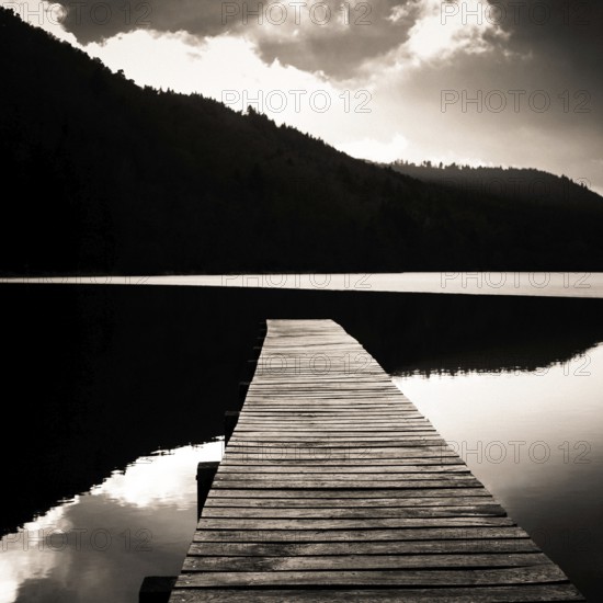 A tranquil wooden pier stretches out over the calm waters of Lake Chambon, surrounded by lush hills in Auvergne, France. This idyllic setting invites peace and relaxation, Puy de Dome, Auvergne Rhone Alpes, France