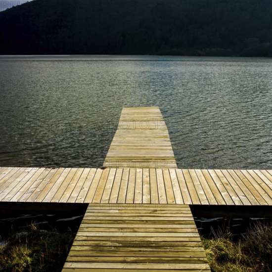A wooden dock reaches out over serene waters reflecting nearby hills, providing a peaceful spot for nature appreciation on a quiet day, lake Chambon, Puy de Dome, Auvergne Rhone Alpes, France
