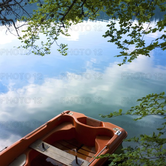 A small boat rests quietly on the tranquil waters of Lake Pavin, surrounded by lush greenery. The clear reflections of clouds and trees create a serene atmosphere of natural beauty, Puy de Dome, Auvergne Rhone Alpes, France