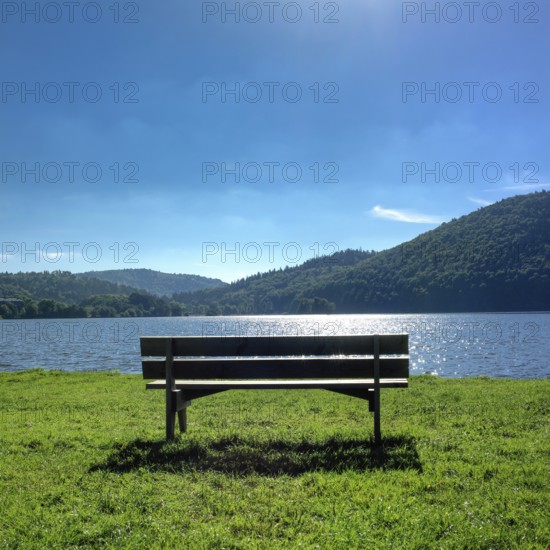 A solitary bench sits on lush green grass by a calm lake in Auvergne, France. The sun shines brightly above, creating a peaceful and reflective atmosphere. Puy de Dome. Auvergne Rhone Alpes. France