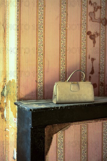 A lonely handbag sits on a worn shelf in an abandoned house, surrounded by peeling wallpaper. The atmosphere reflects neglect and time's passage, evoking nostalgia