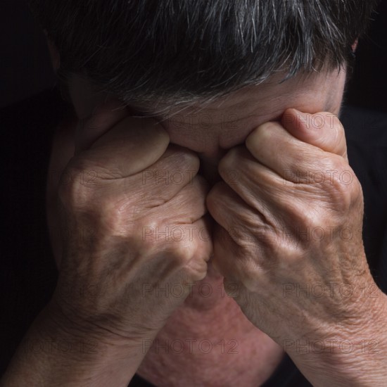 A person sits with their head bowed, hands clasped tightly over their eyes, conveying deep emotion and turmoil. The setting is dark, emphasizing feelings of distress and introspection