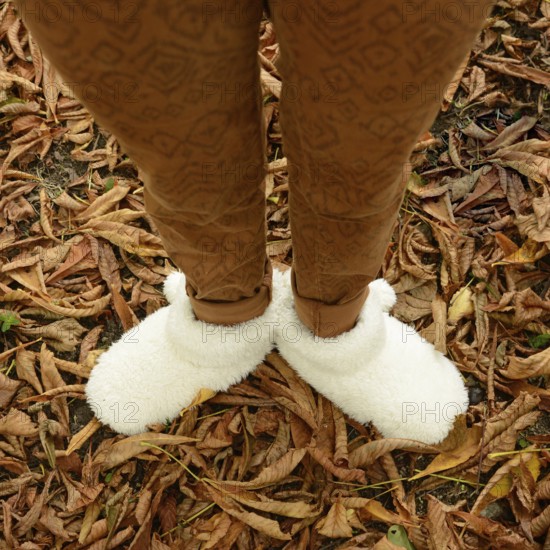 A young woman stands among fallen leaves, wearing warm, fluffy shoes that provide comfort. The autumn landscape reflects vibrant colors, enhancing the cozy atmosphere of the season